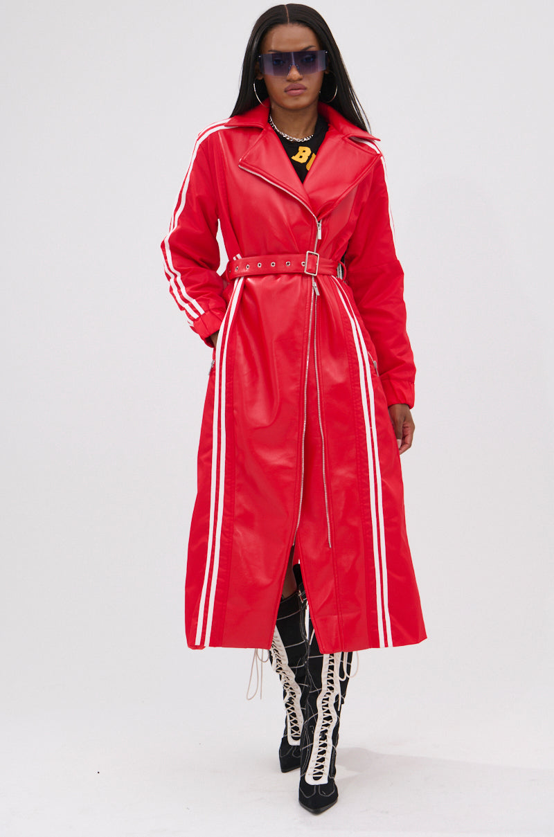 A woman in the UNAVAILABLE MIXED MEDIA TRENCH—a long red belted faux leather coat with white sleeve and front stripes—pairs it with black lace-up boots, dark sunglasses, and a chunky gold necklace for a sporty look against a white backdrop.