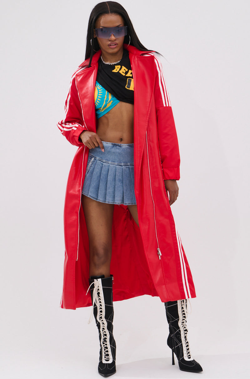 A woman wearing the UNAVAILABLE MIXED MEDIA TRENCH, a black crop top, blue denim pleated mini skirt, black and white lace-up boots, sunglasses, and hoop earrings poses confidently against a plain background.