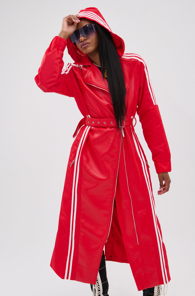 A person wears the UNAVAILABLE MIXED MEDIA TRENCH, a long red faux leather hooded coat with white stripes, belted waist, and zipper front. They adjust the hood, wear blue sunglasses, have long hair, and stand before a plain light gray background.