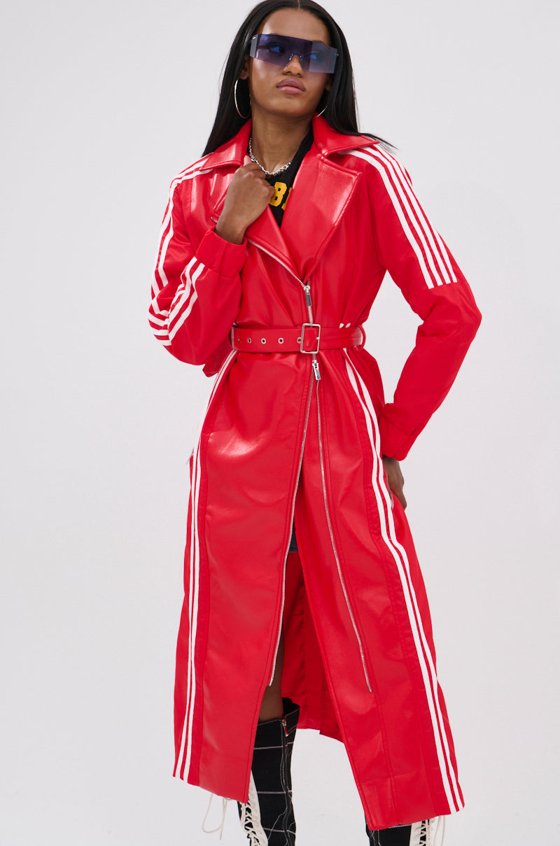 A person models the UNAVAILABLE MIXED MEDIA TRENCH, a long bright red faux leather belted coat with white sleeve and side stripes, styled with large sunglasses, hoop earrings, and tall lace-up boots against a plain background.