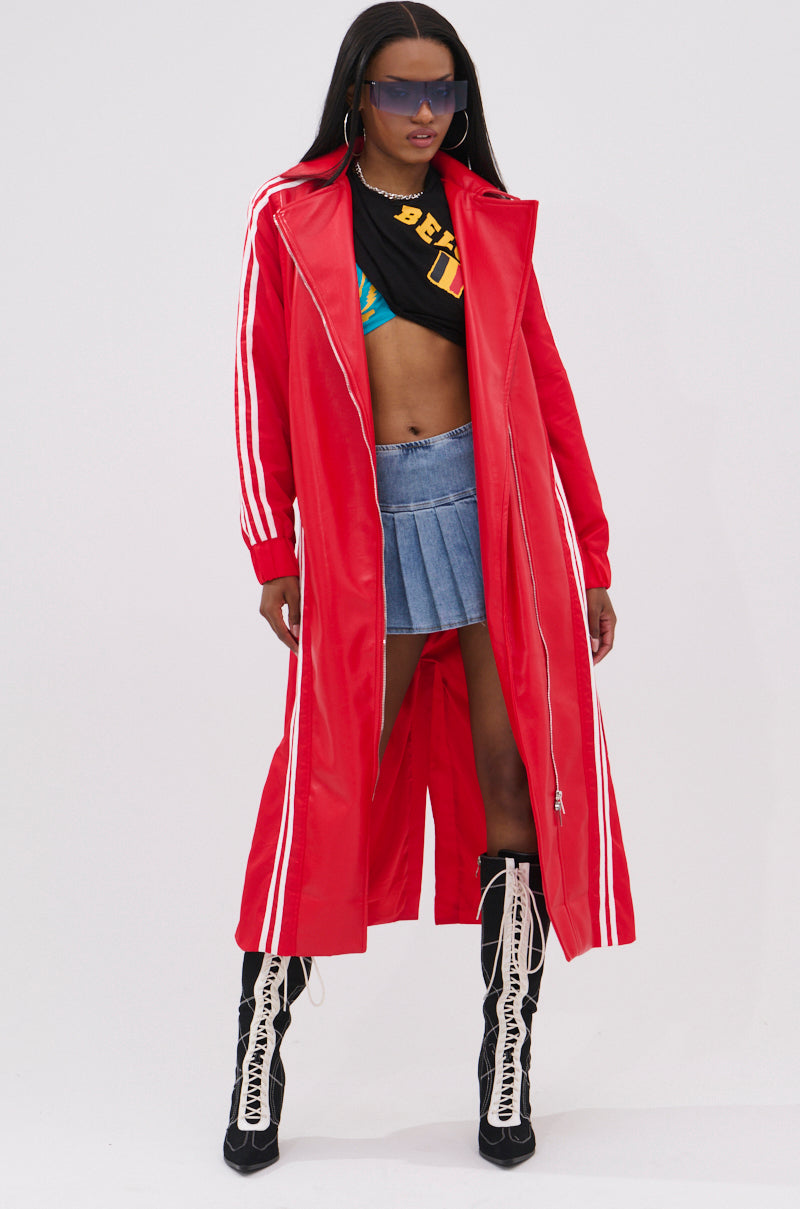 A woman stands confidently in the UNAVAILABLE MIXED MEDIA TRENCH, a long red coat with white stripes, paired with a black crop top, blue mini skirt, tall sporty lace-up boots, and oversized sunglasses against a plain background.