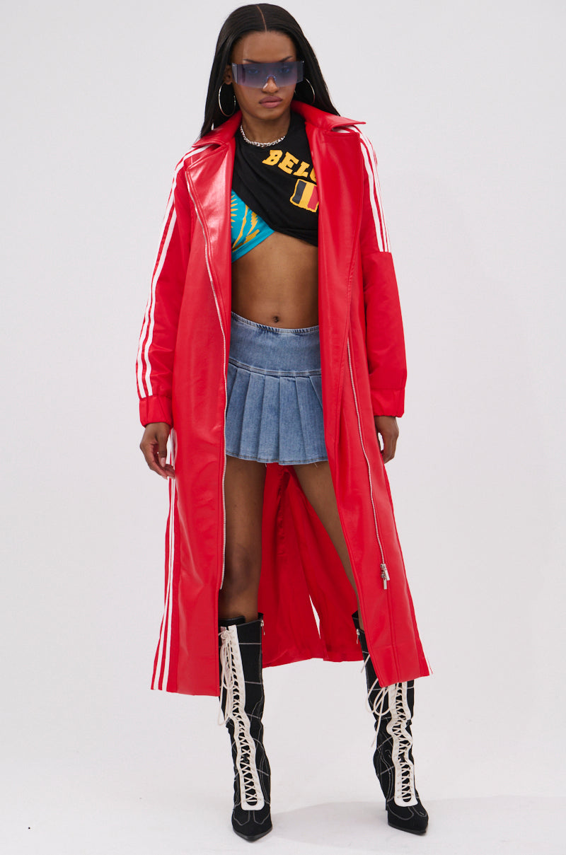 A woman wearing the UNAVAILABLE MIXED MEDIA TRENCH, featuring a long red faux leather design with white stripes, styled with a black crop top over a blue bikini top, mini denim skirt, lace-up boots, and large sunglasses.