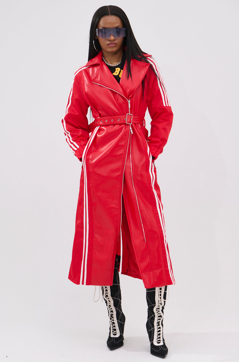 A person wears the UNAVAILABLE MIXED MEDIA TRENCH, featuring a long red belted design with white sleeve stripes, paired with black and white tall lace-up boots, oversized sunglasses, and a chunky gold necklace against a plain light background.