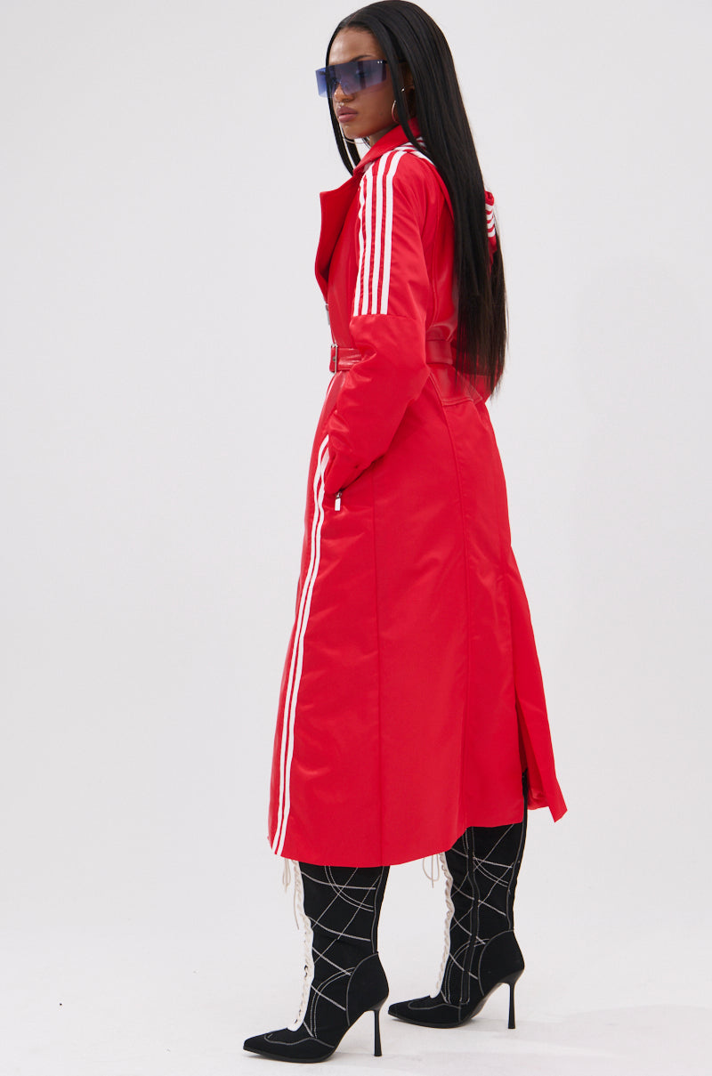 A woman stands sideways in the UNAVAILABLE MIXED MEDIA TRENCH, a long red faux leather coat with white stripes on the sleeves and sides. She pairs it with black high-heeled boots featuring a white grid pattern, blue sunglasses, and straight long hair.