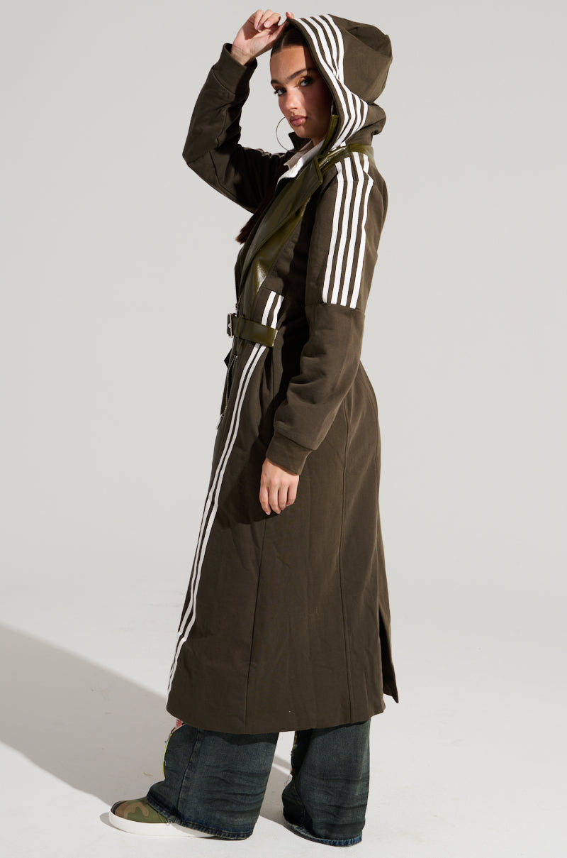 A person in a long HADLEY MIXED MEDIA TRENCH with olive-green and white stripes poses against a plain backdrop. They sport dark wide-leg pants and sneakers, holding the hood with one hand while gazing at the camera. The coat, complete with a belt, offers a compact design that epitomizes Street Style Fashion.