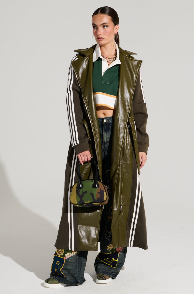 A person with tied-back hair wears the HADLEY MIXED MEDIA TRENCH, a knee-length dark olive-green faux leather coat with white stripes. They pair it with a green and white cropped top and baggy dark jeans featuring patches. Completing the street style look, they hold a small camouflage-patterned handbag against a simple, light-colored background.
