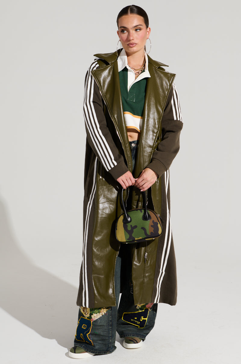 A model stands against a plain backdrop, wearing the HADLEY MIXED MEDIA TRENCH—a long, olive green faux leather coat with white stripes on the sleeves—paired with a green and white rugby shirt and baggy jeans adorned with patches. She holds a small camouflage bag, sports slicked-back hair, and minimalistic makeup, perfectly embodying street style fashion.