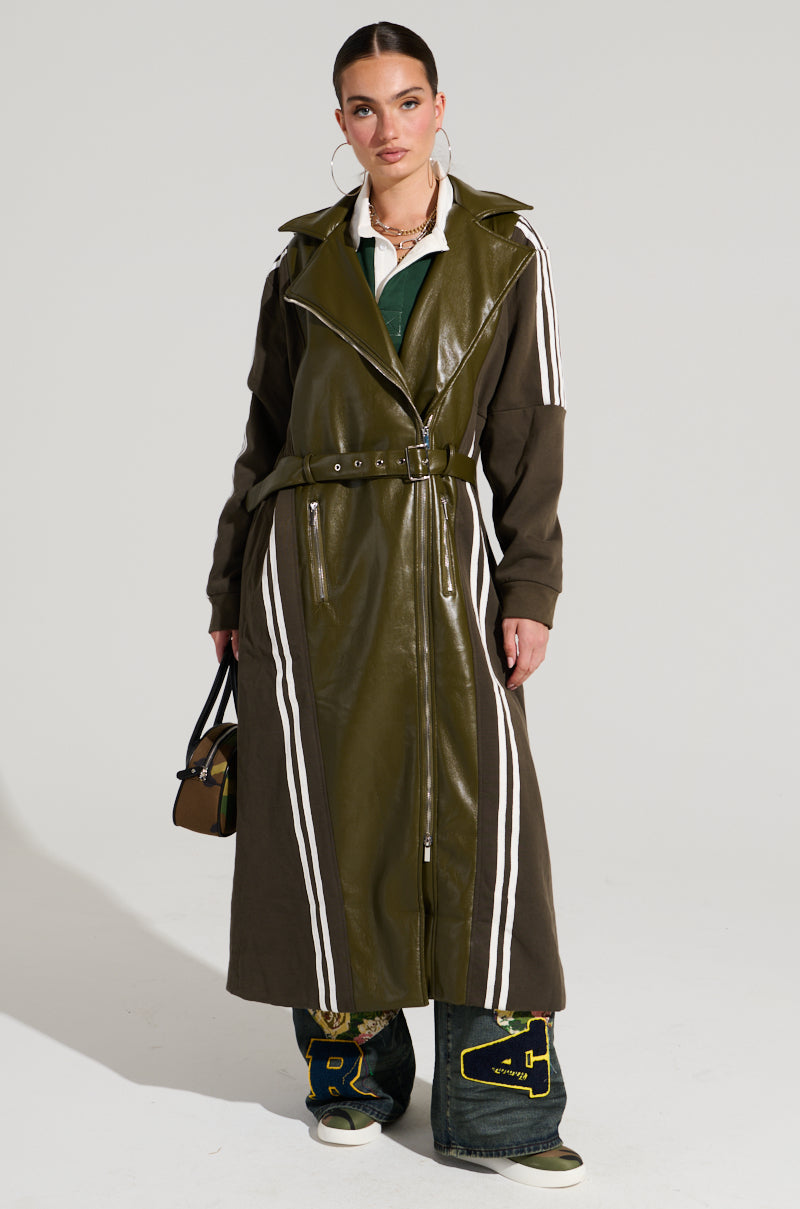 A model wearing the HADLEY MIXED MEDIA TRENCH, which features white stripes on the sleeves and sides. The faux leather coat with a belted waist is paired perfectly with wide-leg jeans adorned with large, colorful patches and intricate embroidery. Embracing street style fashion, the model completes the look by holding a small brown handbag.