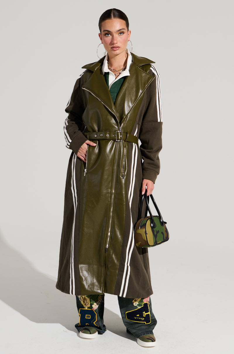A person stands confidently against a plain background, embodying Street Style Fashion in the HADLEY MIXED MEDIA TRENCH, an olive green trench coat with white stripes. Their layered outfit includes a green top and embroidered pants. They carry a small camouflage-patterned handbag and accessorize with large hoop earrings.