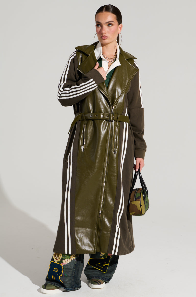 A model stands wearing the HADLEY MIXED MEDIA TRENCH in olive green with white stripes on the sleeves and sides, complemented by wide blue jeans adorned with embroidered patches and white sneakers. They carry a small camo bag and accessorize their slicked-back hair with layered necklaces, perfectly epitomizing street style fashion.
