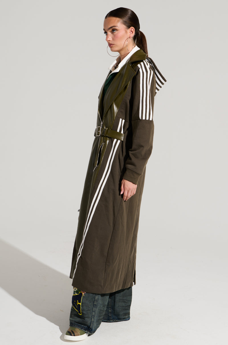 A person with straight, dark hair tied back wears the HADLEY MIXED MEDIA TRENCH, a long olive-green coat featuring white stripes along the sleeves and back, cinched at the waist with a belt. They are also wearing wide-legged dark jeans and have a serious expression while looking sideways, embodying street style fashion.