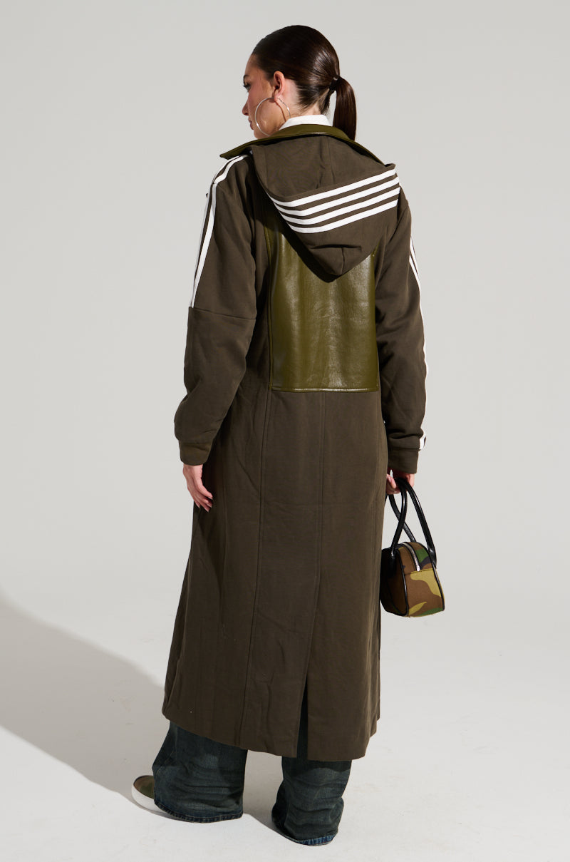 A person with long dark hair pulled back in a ponytail is wearing the HADLEY MIXED MEDIA TRENCH, a long, dark green coat featuring white stripes on the shoulders and an olive green panel on the back. They're also holding a small camouflage-patterned handbag while facing away from the camera, exemplifying street style fashion.