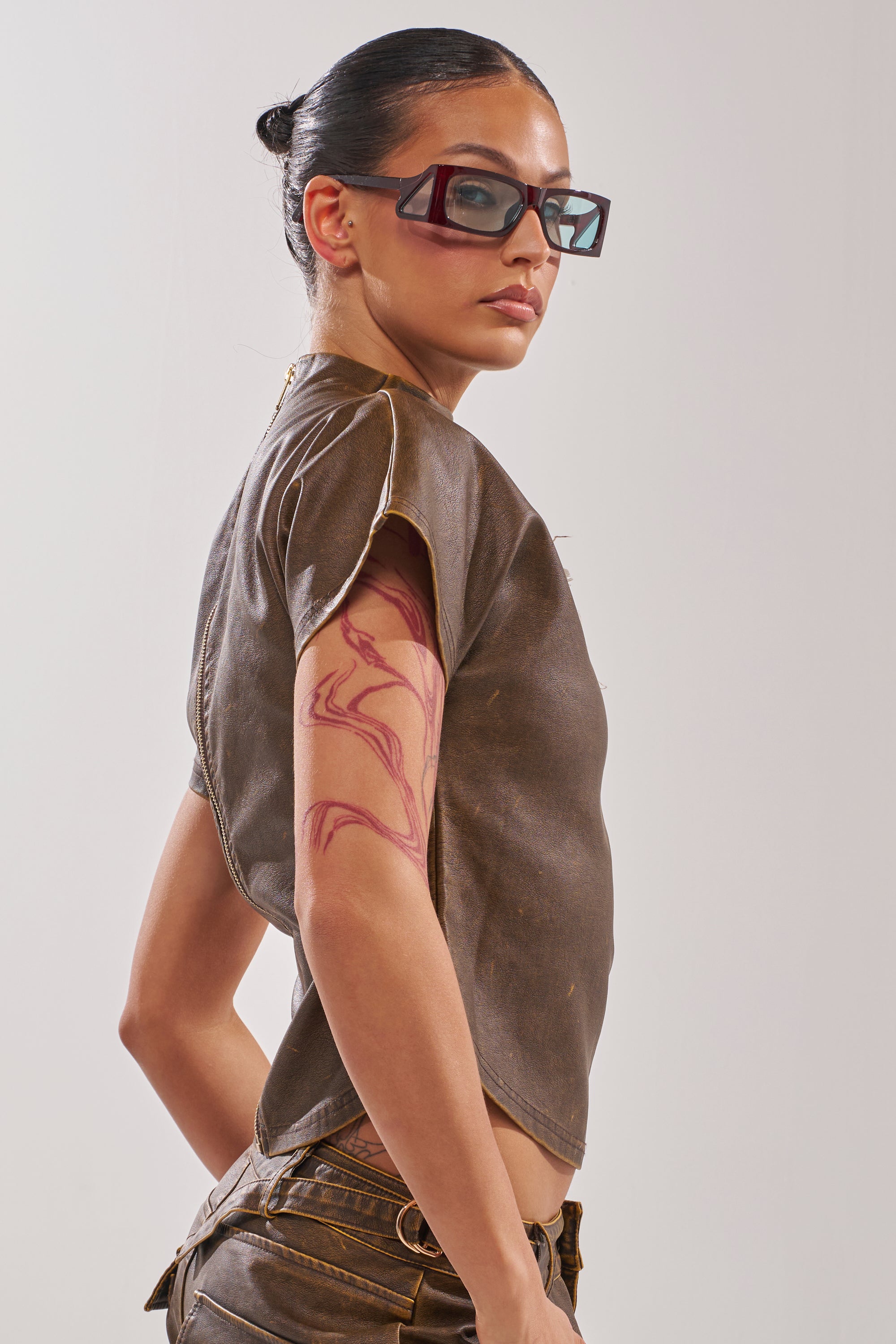 A person with slicked-back dark hair and angular sunglasses glances over their shoulder, wearing the ALL AROUND THE CITY MOCK NECK TOP, which reveals an abstract upper arm tattoo against a plain, light background. 3