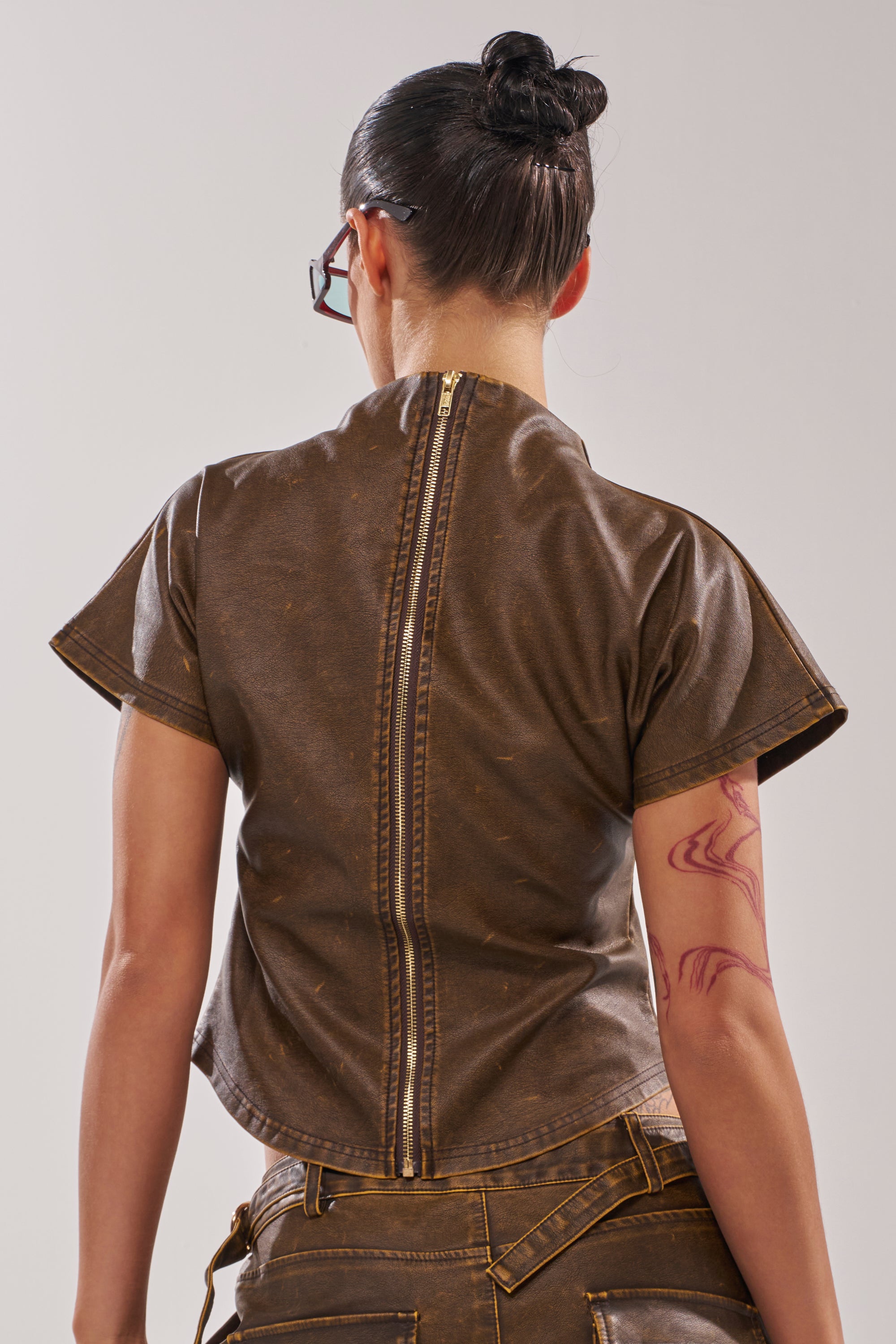 A person with dark hair in a bun and glasses stands with their back to the camera, wearing the ALL AROUND THE CITY MOCK NECK TOP featuring a faux leather finish and visible back zipper. A red abstract tattoo is on their left upper arm. 4