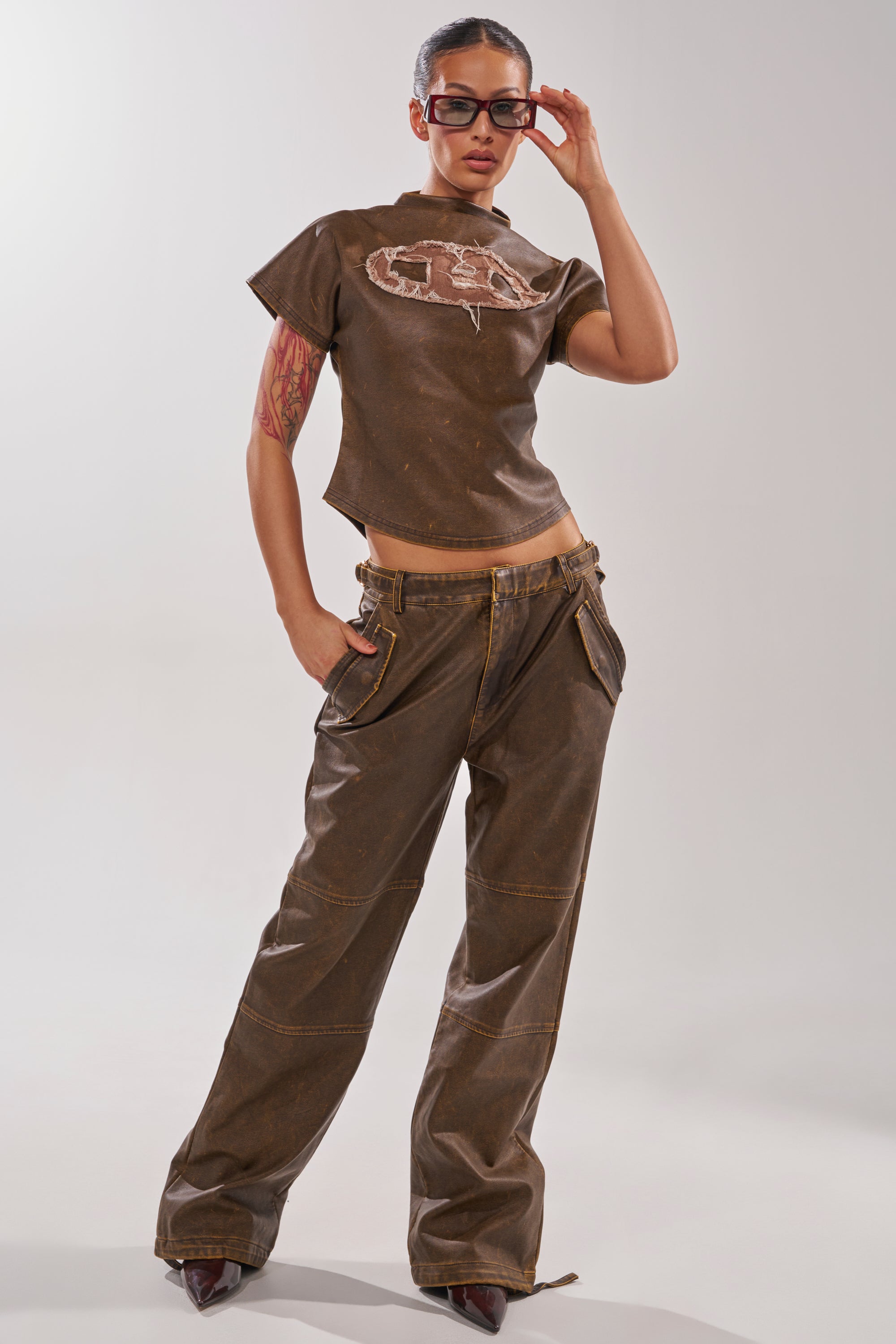 A woman poses confidently in brown wide-leg pants and the ALL AROUND THE CITY MOCK NECK TOP with a graphic design, paired with sunglasses and maroon pointed shoes, against a plain light background. 5
