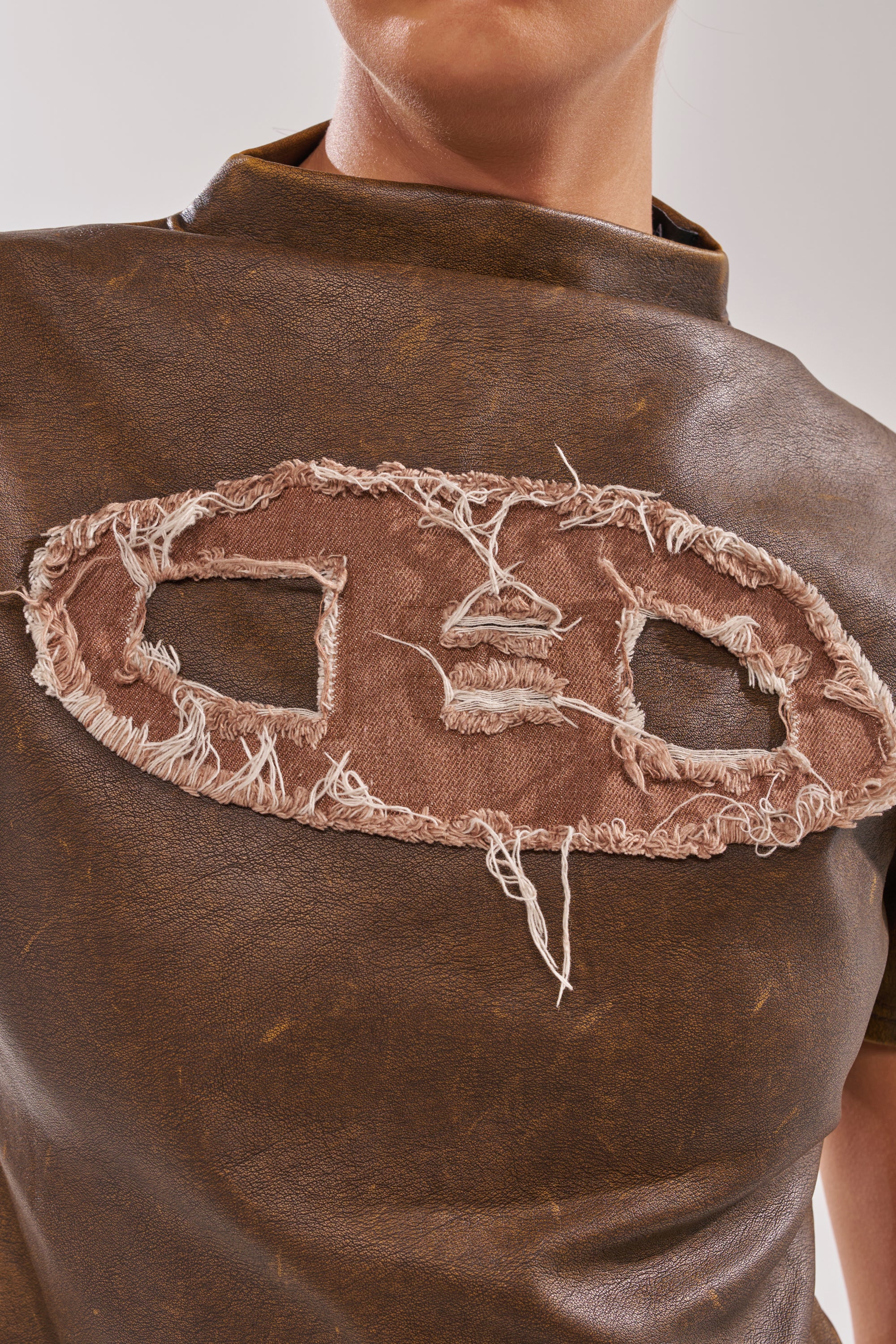 A close-up of a person wearing the ALL AROUND THE CITY MOCK NECK TOP in brown faux leather, featuring a prominent oval frayed patch with two rectangular cutouts and rough stitching on the chest. 8