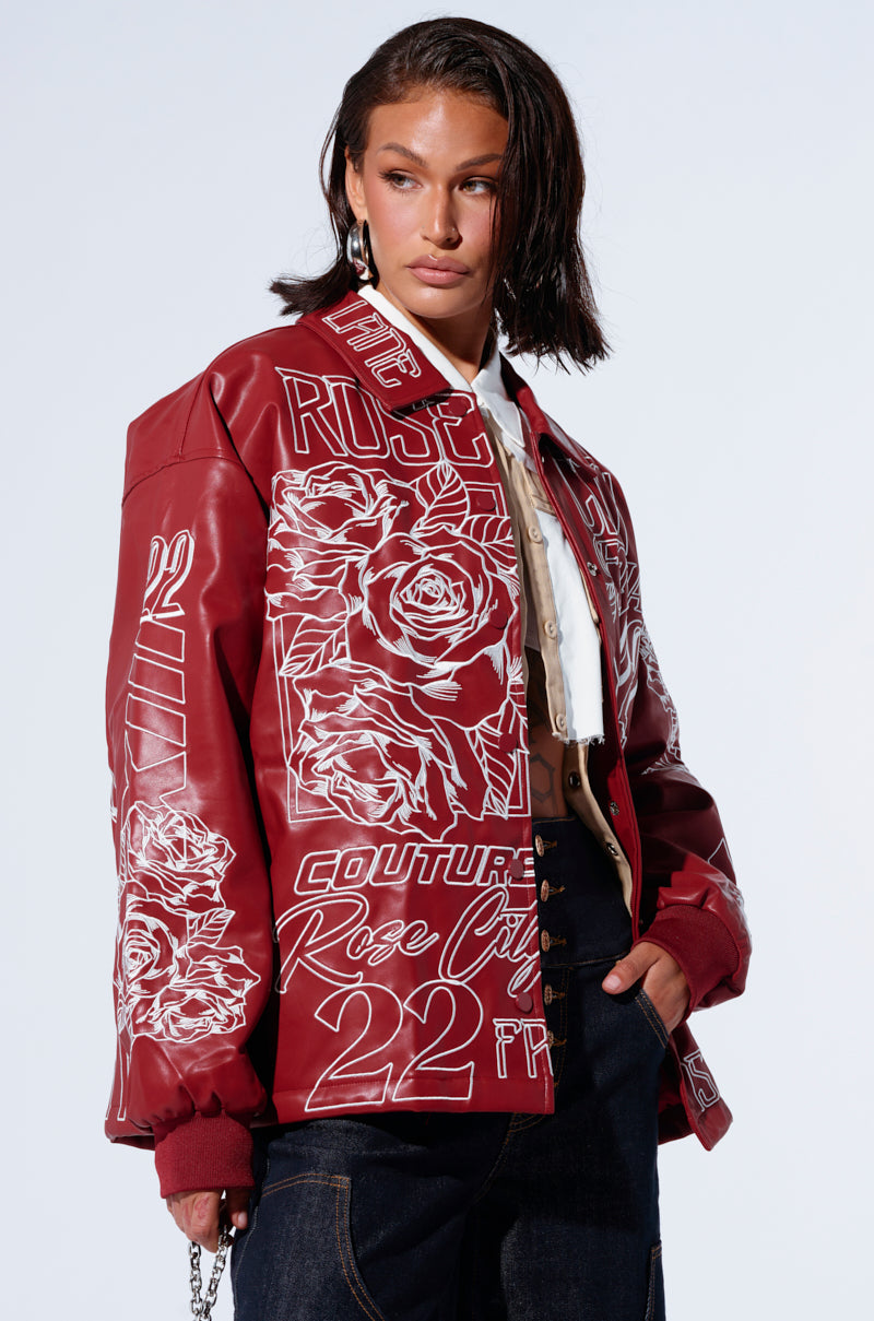 A person with shoulder-length brown hair confidently models the ROSE CITY EMBROIDERED BOMBER, a red faux leather jacket adorned with white rose graphics and text. They wear a white shirt underneath and dark jeans, accessorized with earrings and rings, standing against a light grey background.
