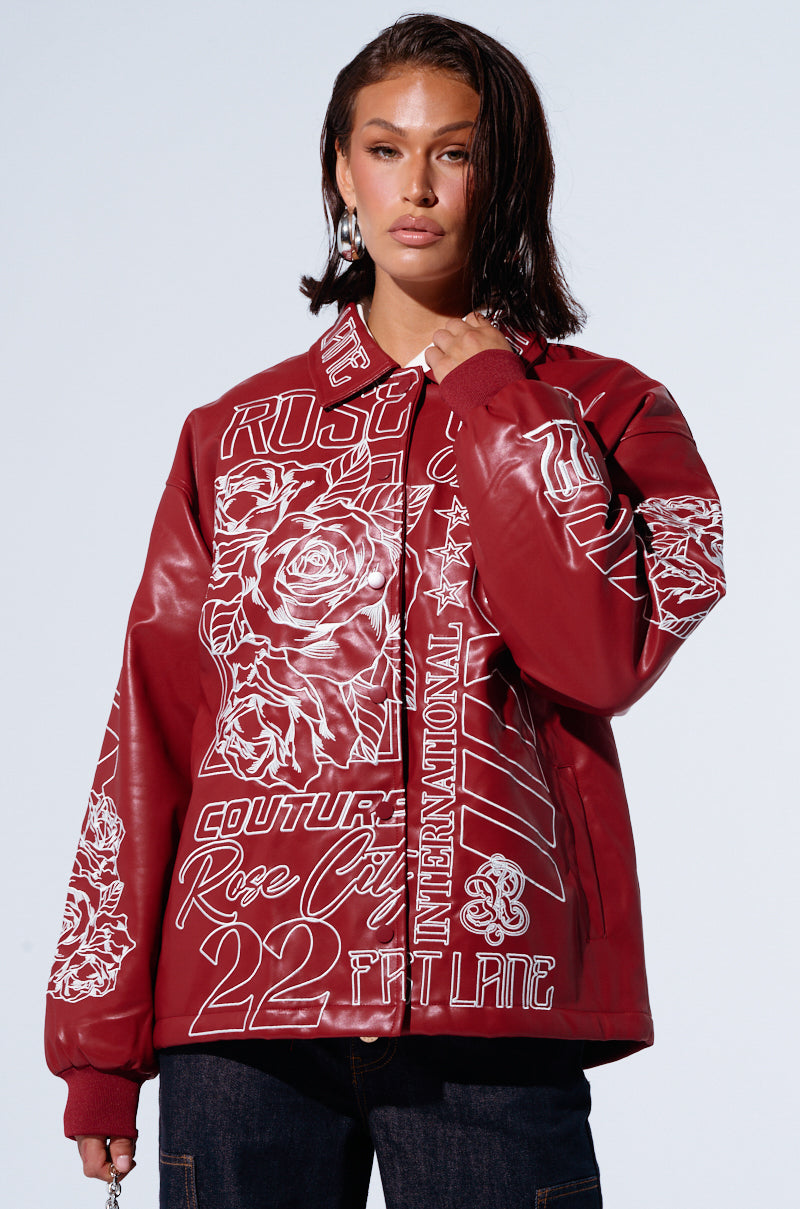 A person with shoulder-length dark hair is wearing an oversized ROSE CITY EMBROIDERED BOMBER, decorated with various white graphic designs such as roses, text, and numbers. The faux leather jacket is zipped up as the person stands against a plain, light-colored background.