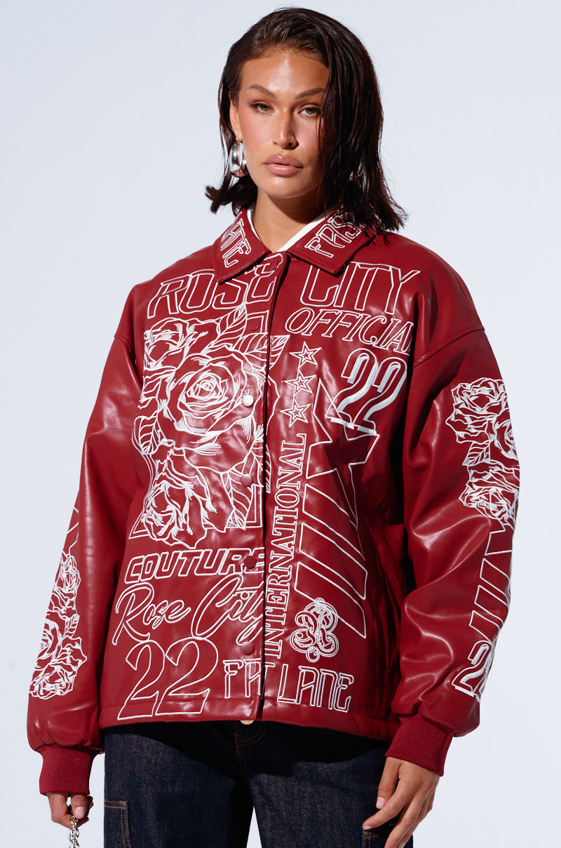 A person with shoulder-length dark hair wears the ROSE CITY EMBROIDERED BOMBER in maroon, featuring intricate white graphic designs of roses, numbers, and various text phrases. The oversized silhouette pairs stylishly with dark pants as the wearer stands against a light background.