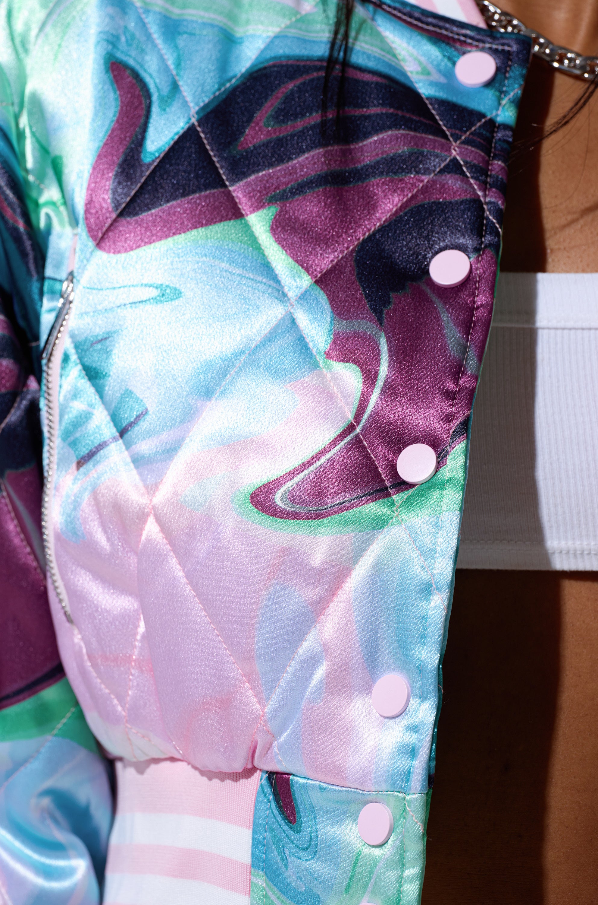 Close-up of the HEART TO HEART SATIN BOMBER, a quilted pastel jacket with abstract blue, pink, and purple swirls and light pink buttons. The cropped bomber is styled over a white top, revealing a hint of silver necklace and bare skin.