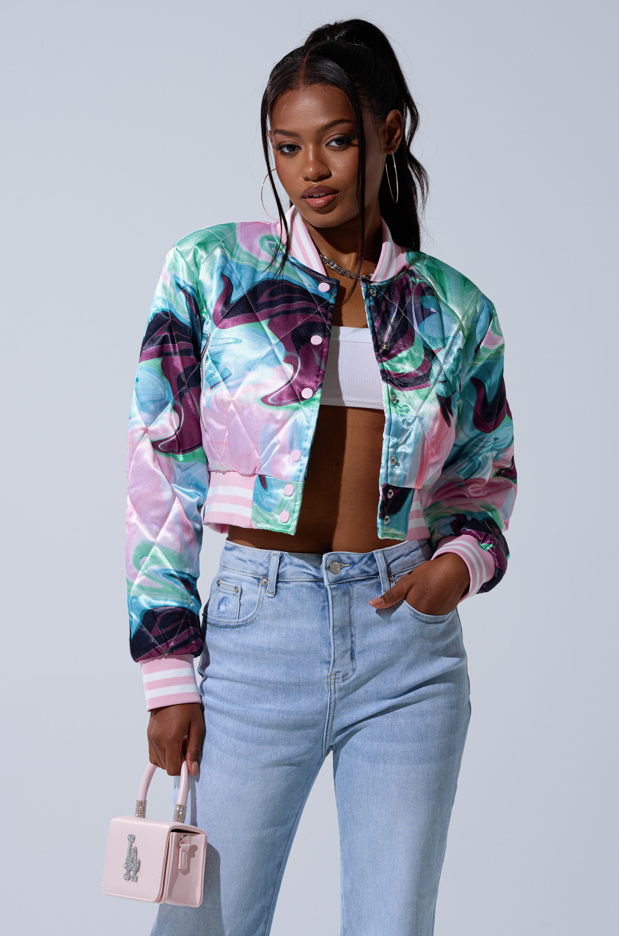 A woman stands confidently in the HEART TO HEART SATIN BOMBER, a vibrant cropped jacket ideal for pastel outerwear, paired with a white top, light blue high-waisted jeans, and a small pale pink handbag. Her hair is in a high ponytail.