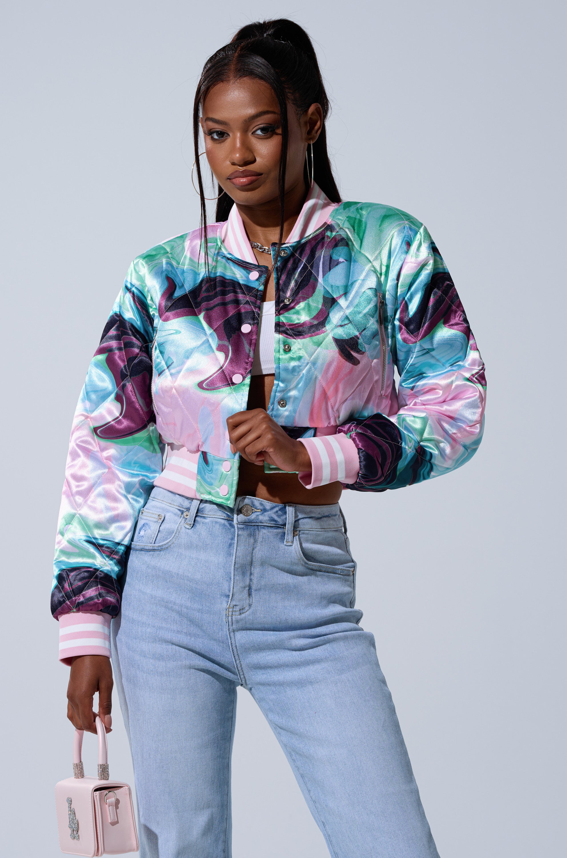 A woman wearing the HEART TO HEART SATIN BOMBER with pastel swirls, light blue high-waisted jeans, and a small pink purse poses confidently against a plain light background.
