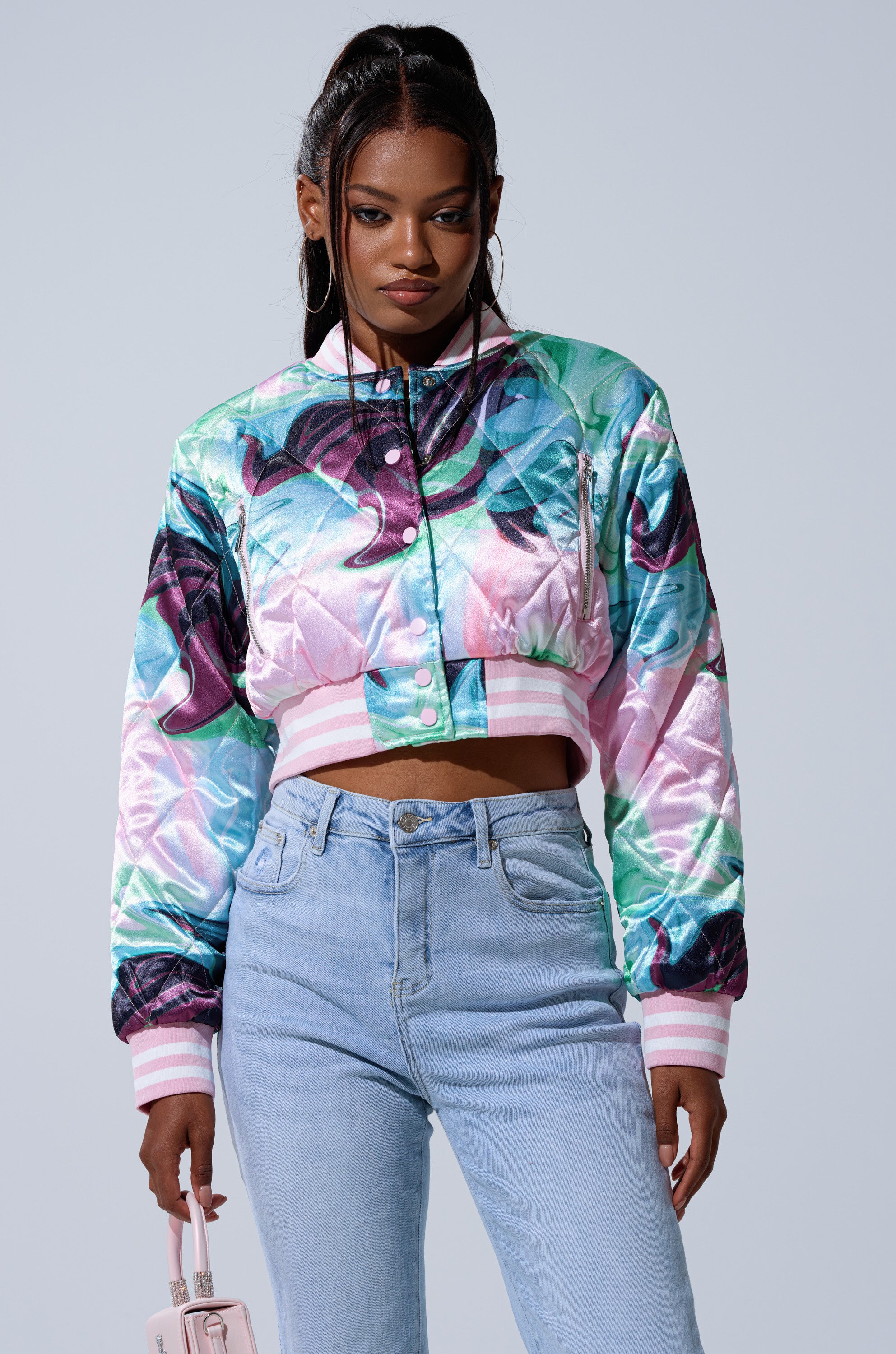 A woman wearing the HEART TO HEART SATIN BOMBER with a colorful floral pattern and high-waisted light blue jeans stands against a plain background, holding a small pale pink handbag.