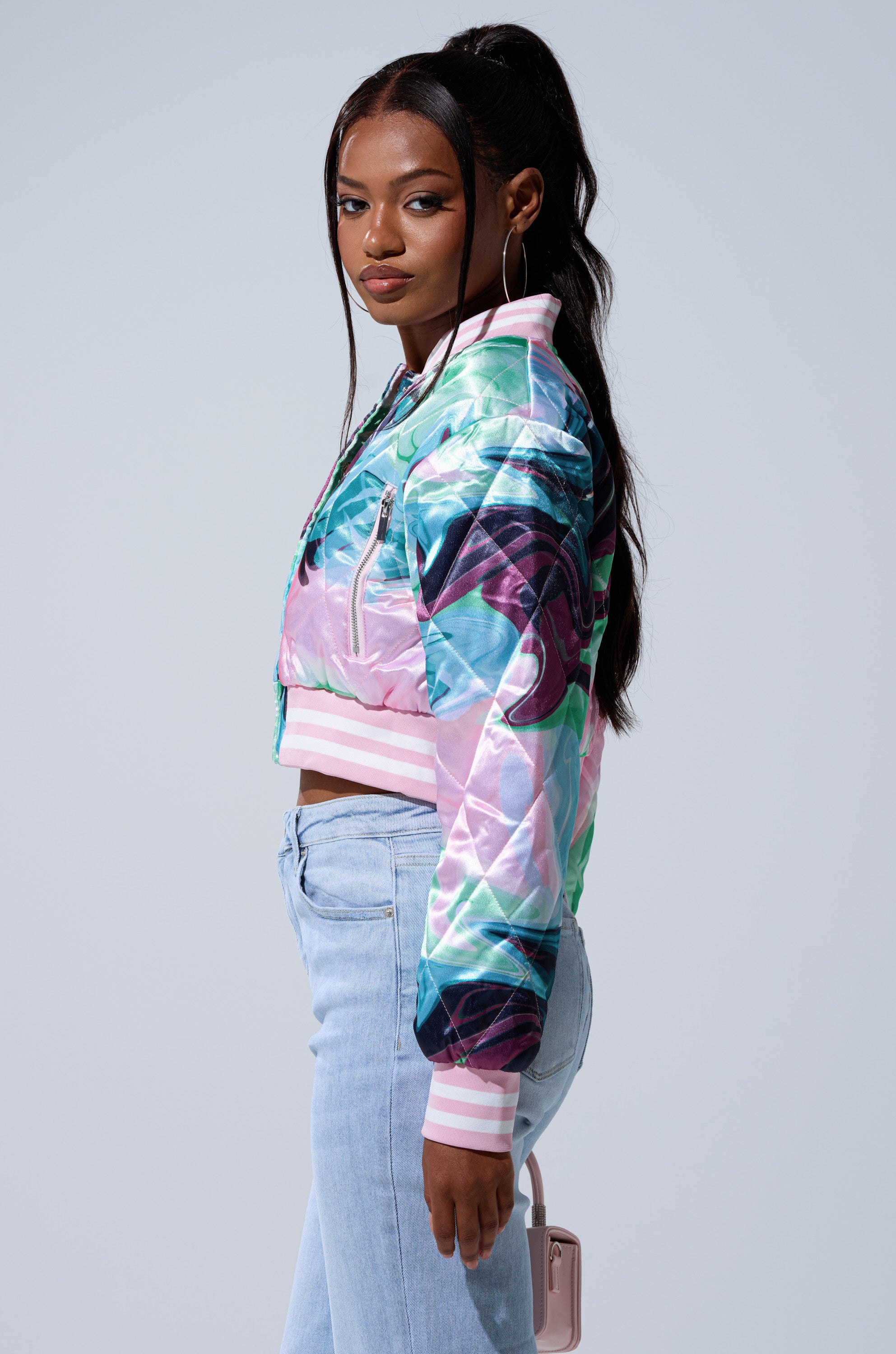 A woman with long dark hair in a high ponytail wears the HEART TO HEART SATIN BOMBER, styled with light blue jeans and hoop earrings. She stands sideways, holding a light pink purse against a plain background.
