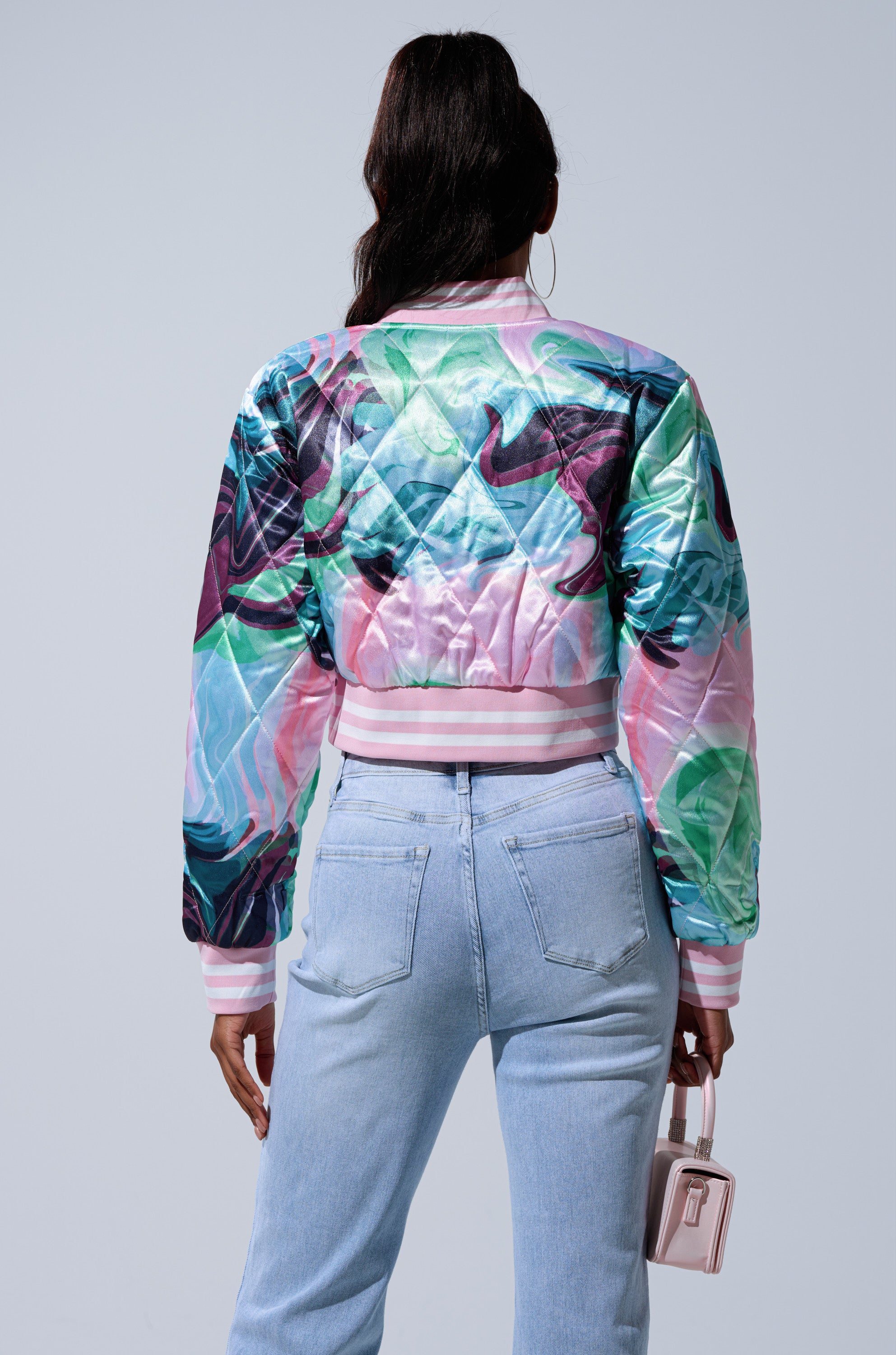 A person stands facing away, wearing the HEART TO HEART SATIN BOMBER with abstract pastel patterns, light blue high-waisted jeans, and holding a small pink handbag against a plain light background.