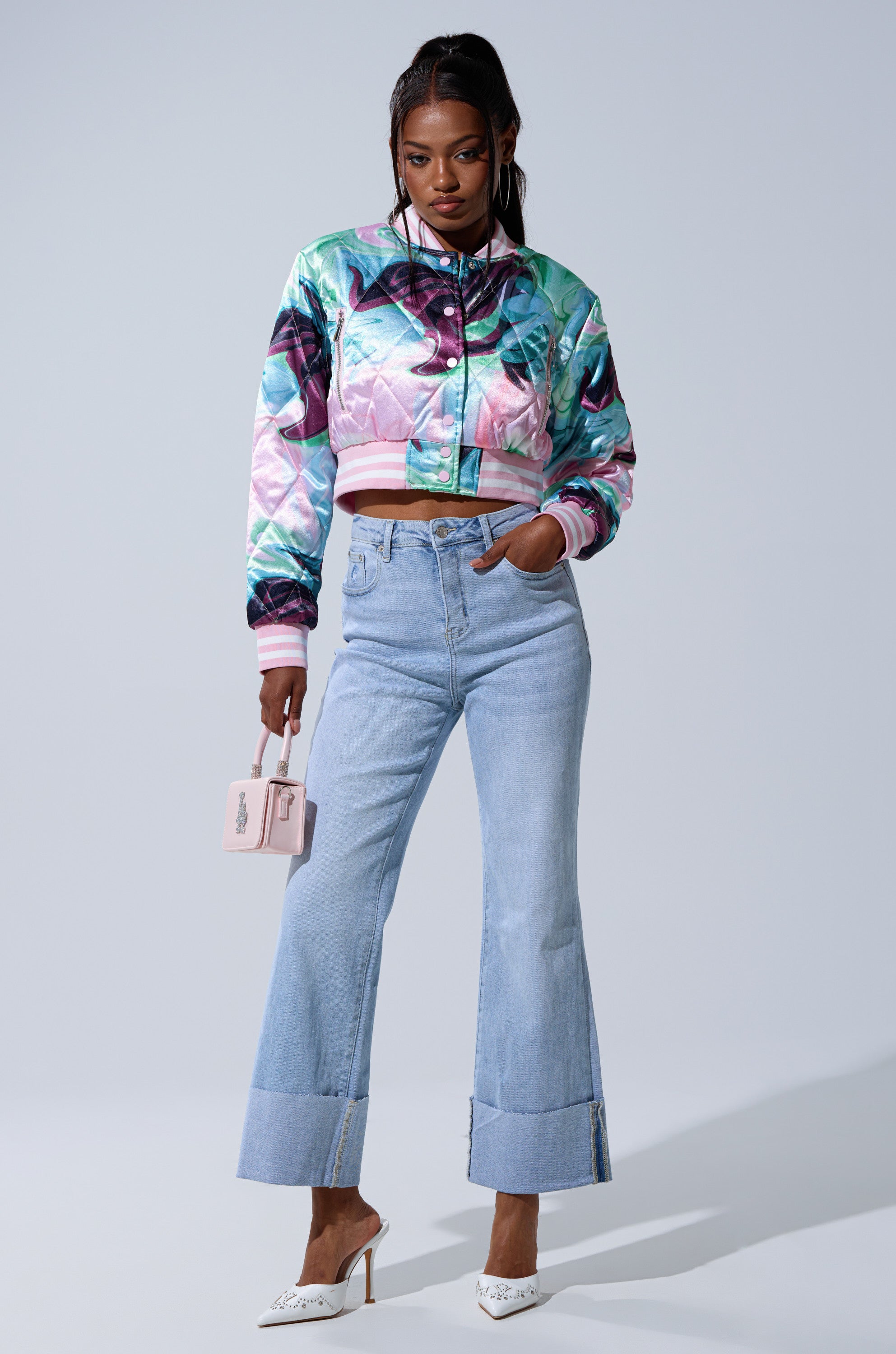 A woman stands against a plain background wearing the colorful HEART TO HEART SATIN BOMBER, high-waisted light blue flared jeans with wide cuffs, white heels, and holding a small light pink handbag.