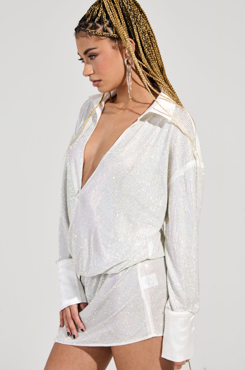 A woman with braided hair poses confidently in profile, wearing the LOGEN RHINESTONE MINI DRESS, a sparkly silver outfit embellished with sequins. The dress features a deep V-neck and she accessorizes her party look with long earrings against a plain background.