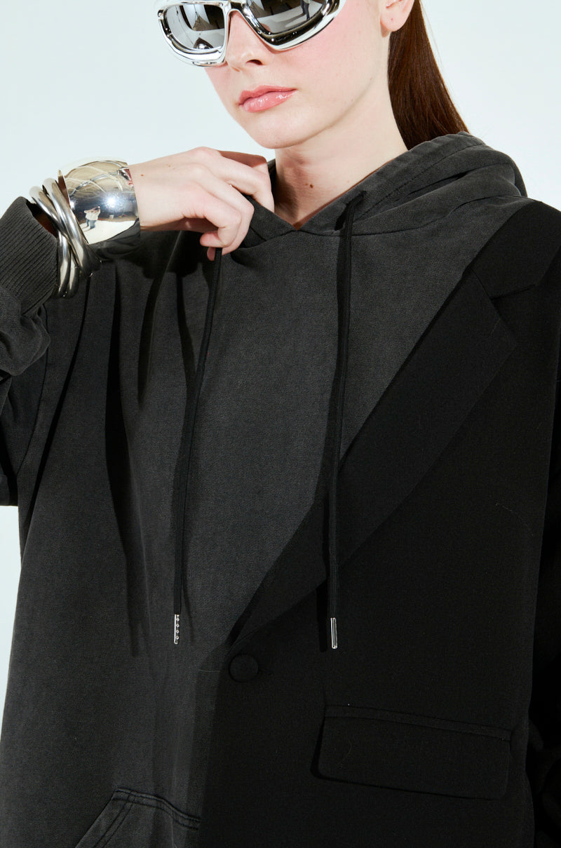 Wearing futuristic sunglasses, a black hoodie, and the CASUAL BUSINESS SWEATSHIRT BLAZER MINI DRESS, a person holds the hoodie collar. Their left wrist is adorned with metallic bracelets against an off-white background.