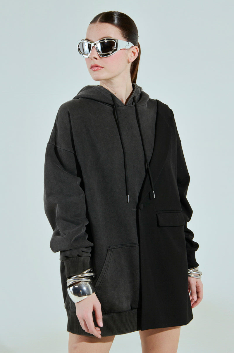 A person in oversized sunglasses and a gray cotton-blend hoodie with a black jacket overlay, their hair in a sleek ponytail, wears a large metallic bracelet. The plain light background enhances the CASUAL BUSINESS SWEATSHIRT BLAZER MINI DRESS vibe.