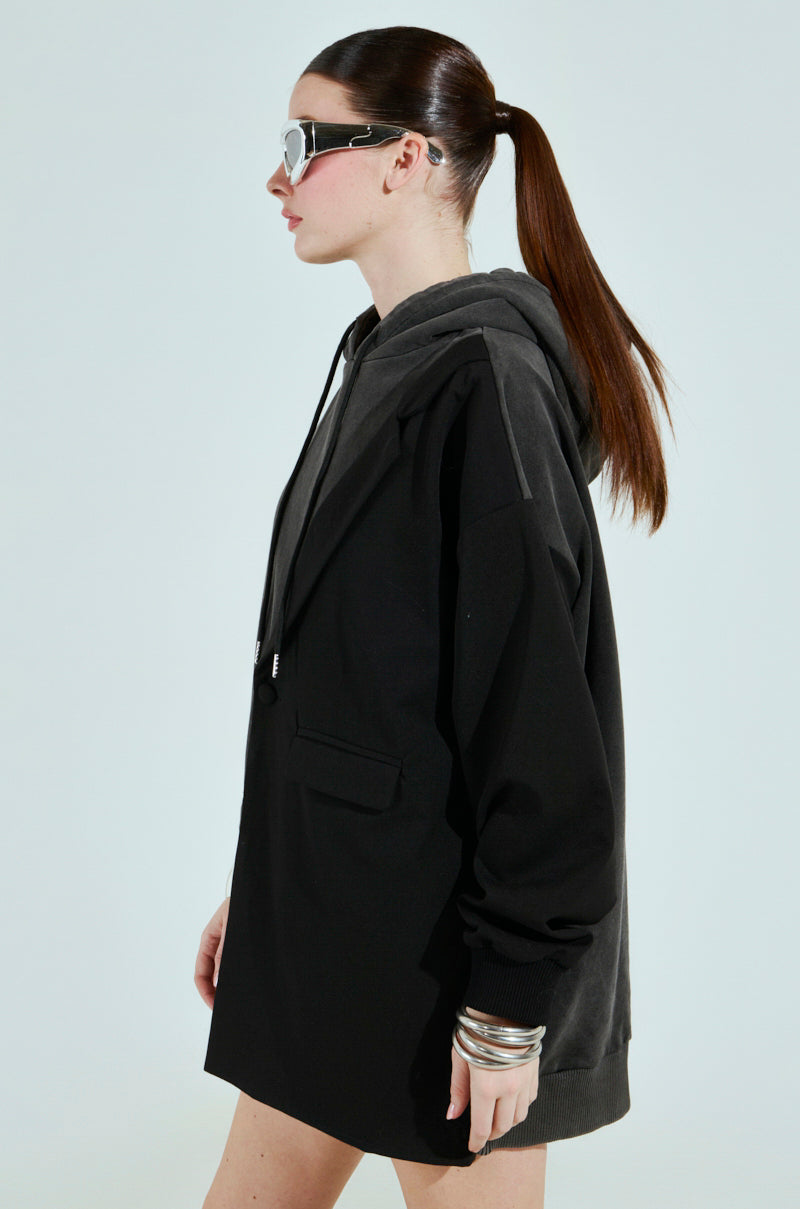 A person with long hair in a ponytail is wearing an oversized, black cotton-blend CASUAL BUSINESS SWEATSHIRT BLAZER MINI DRESS over a hoodie and large sunglasses. They have multiple bangles on one wrist and are standing against a plain background, facing sideways.