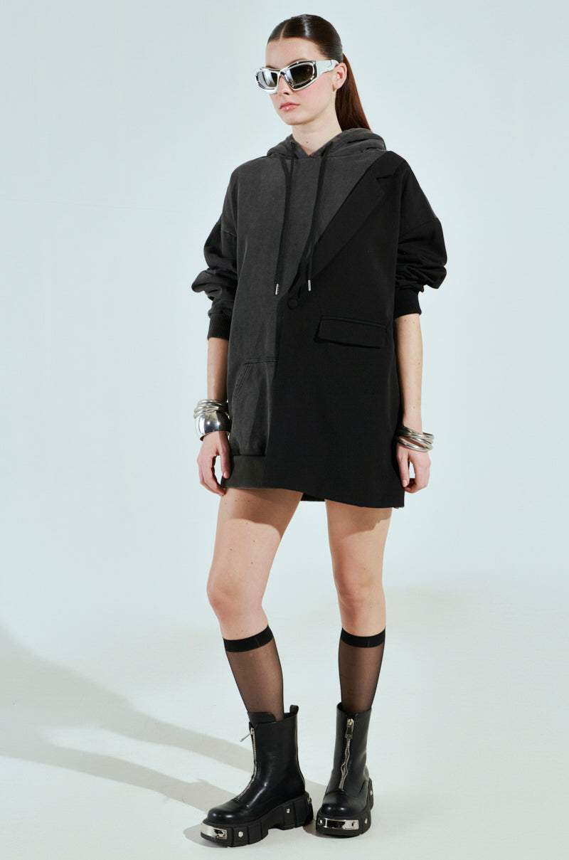 Wearing large sunglasses and chunky silver bracelets, a person confidently models the CASUAL BUSINESS SWEATSHIRT BLAZER MINI DRESS—a half black, half gray cotton-blend oversized hoodie. They pair it with knee-high sheer black socks and black boots with metal details against a plain light gray backdrop.
