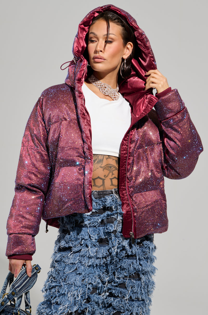 A person wearing the FAYE Rhinestone Puffer in burgundy, which features a hood, layered over a white top that reveals a midriff tattoo. They carry a blue textured handbag and are dressed in a jean skirt with a fringed, layered design.