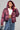 A person wearing the FAYE Rhinestone Puffer in burgundy, which features a hood, layered over a white top that reveals a midriff tattoo. They carry a blue textured handbag and are dressed in a jean skirt with a fringed, layered design.