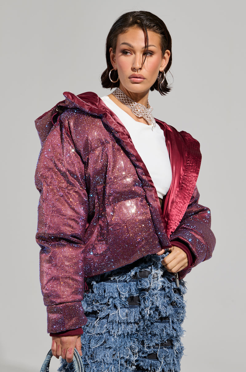 A woman confidently stands, wearing a dazzling magenta jacket over a white top. Her ensemble is enhanced by a chunky necklace, hoop earrings, and a small handbag. She pairs it with a layered, frayed denim skirt that adds an edgy touch to her style as she gazes into the distance, perfectly embodying winter chic without needing the Faye Rhinestone Puffer in Burgundy.