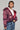 A woman confidently stands, wearing a dazzling magenta jacket over a white top. Her ensemble is enhanced by a chunky necklace, hoop earrings, and a small handbag. She pairs it with a layered, frayed denim skirt that adds an edgy touch to her style as she gazes into the distance, perfectly embodying winter chic without needing the Faye Rhinestone Puffer in Burgundy.