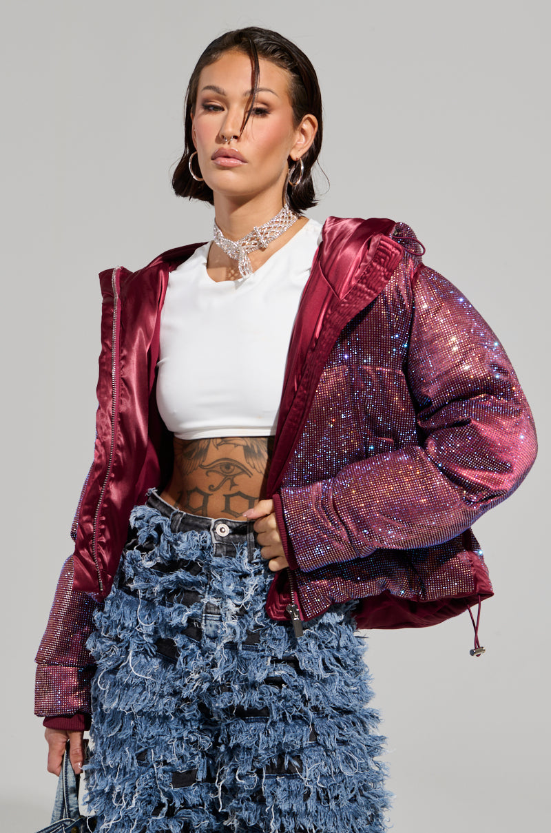A woman poses confidently, wearing a white crop top and the eye-catching FAYE RHINESTONE PUFFER IN BURGUNDY with a hood. Her high-waisted blue denim skirt boasts a textured, frayed design. She completes her look with hoop earrings and a sparkly choker.