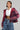 A woman poses confidently, wearing a white crop top and the eye-catching FAYE RHINESTONE PUFFER IN BURGUNDY with a hood. Her high-waisted blue denim skirt boasts a textured, frayed design. She completes her look with hoop earrings and a sparkly choker.