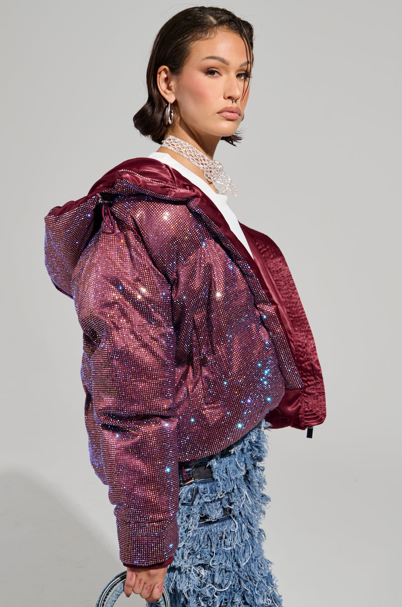A person with short hair is wearing the FAYE Rhinestone Puffer in Burgundy over a white top. They are also adorned with ornate earrings and a choker necklace, paired with blue textured pants. Standing against a plain background, they gaze to the side.