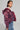 A person with short hair is wearing the FAYE Rhinestone Puffer in Burgundy over a white top. They are also adorned with ornate earrings and a choker necklace, paired with blue textured pants. Standing against a plain background, they gaze to the side.