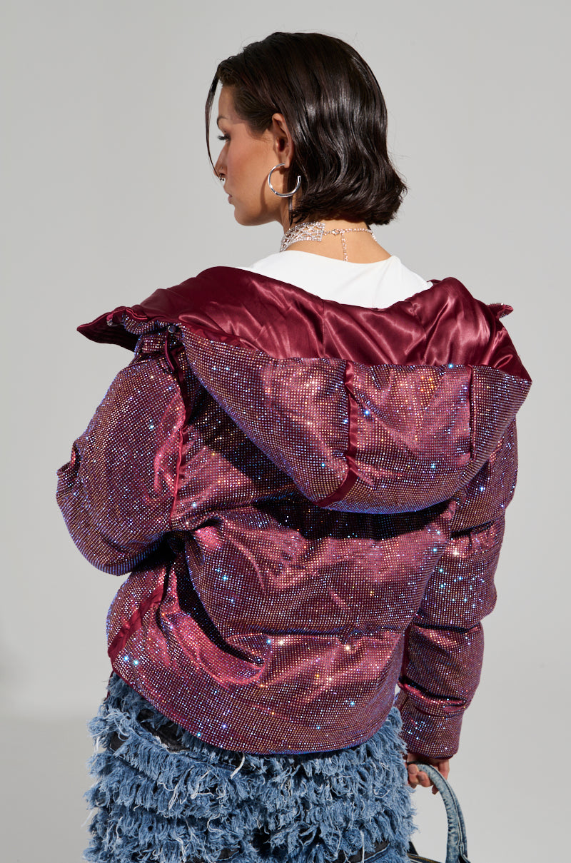 A person with short hair sports the FAYE RHINESTONE PUFFER IN BURGUNDY over a white shirt, paired with a textured, ruffled blue skirt. The setting is a simple, neutral background that exudes the cozy vibe of a winter coat retaining its warm charm.