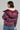 A person with short hair sports the FAYE RHINESTONE PUFFER IN BURGUNDY over a white shirt, paired with a textured, ruffled blue skirt. The setting is a simple, neutral background that exudes the cozy vibe of a winter coat retaining its warm charm.