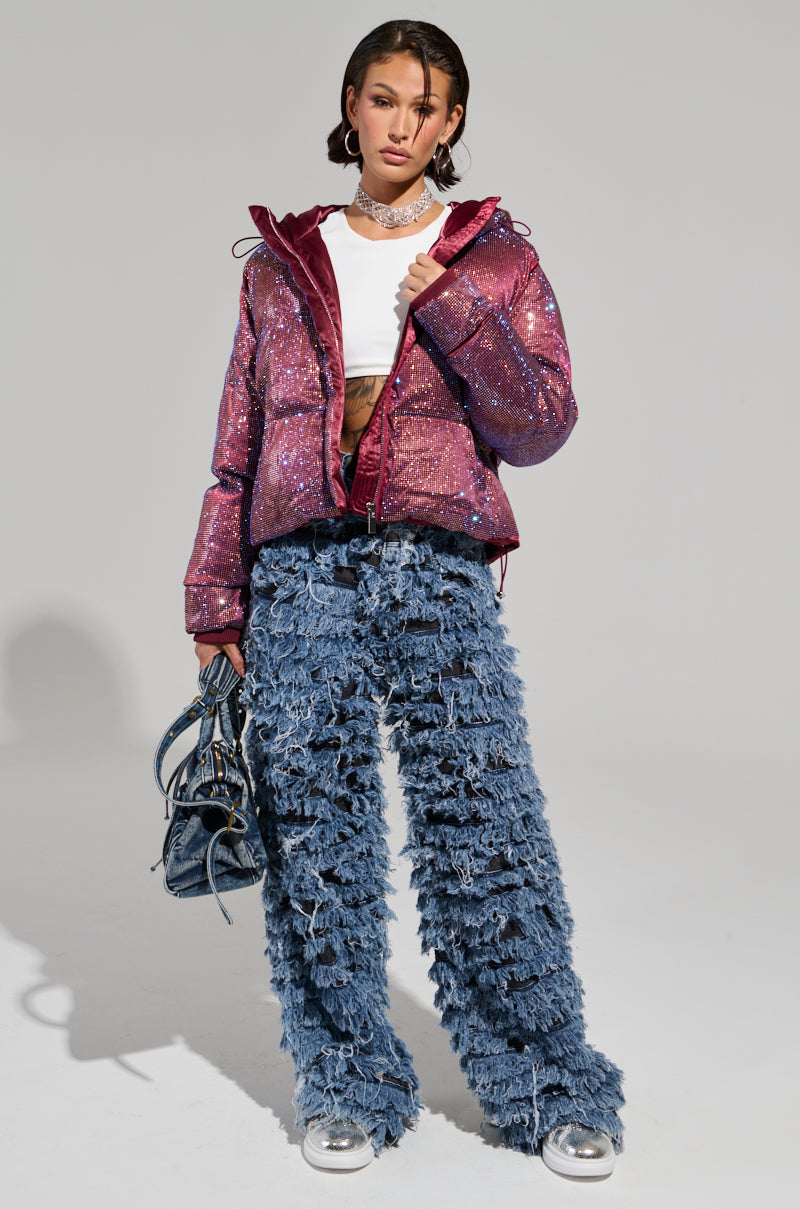 A person poses confidently in the FAYE RHINESTONE PUFFER IN BURGUNDY over a white crop top and textured, fringed blue jeans. They carry a small black handbag and accessorize with a choker necklace and silver sneakers, standing against a plain background.