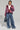 A person poses confidently in the FAYE RHINESTONE PUFFER IN BURGUNDY over a white crop top and textured, fringed blue jeans. They carry a small black handbag and accessorize with a choker necklace and silver sneakers, standing against a plain background.