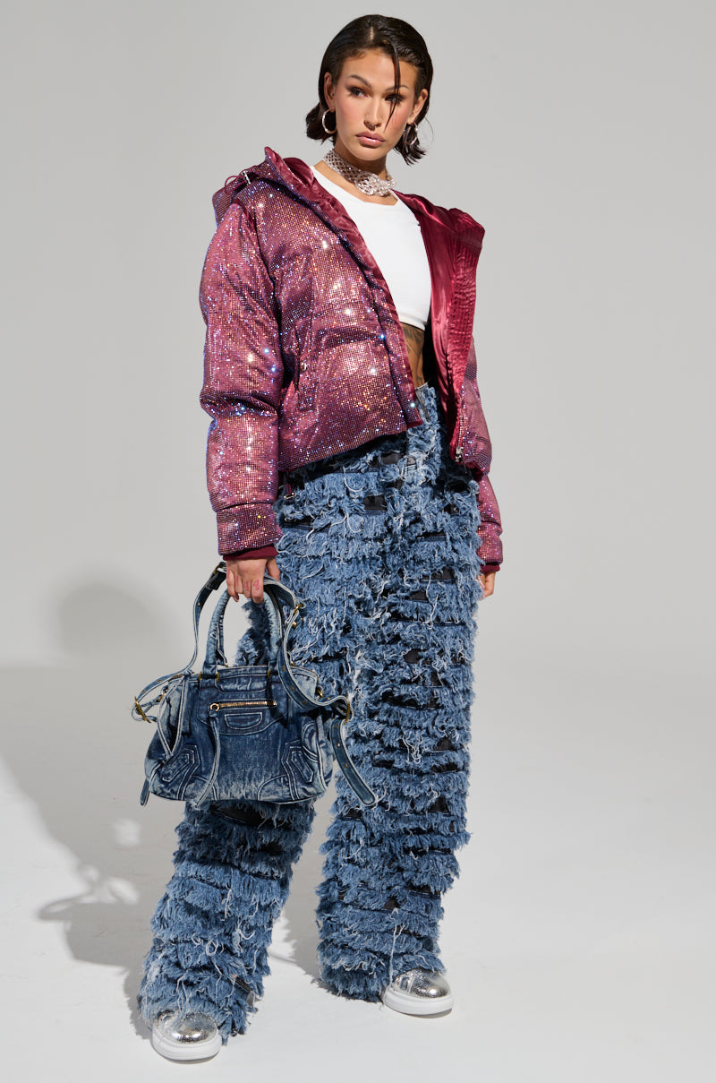 A person poses confidently against a plain background, wearing the FAYE RHINESTONE PUFFER IN BURGUNDY over a white crop top and high-waisted, textured blue pants. They carry a large denim handbag and wear silver shoes, with short, styled hair and minimal accessories.