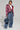A person poses confidently against a plain background, wearing the FAYE RHINESTONE PUFFER IN BURGUNDY over a white crop top and high-waisted, textured blue pants. They carry a large denim handbag and wear silver shoes, with short, styled hair and minimal accessories.