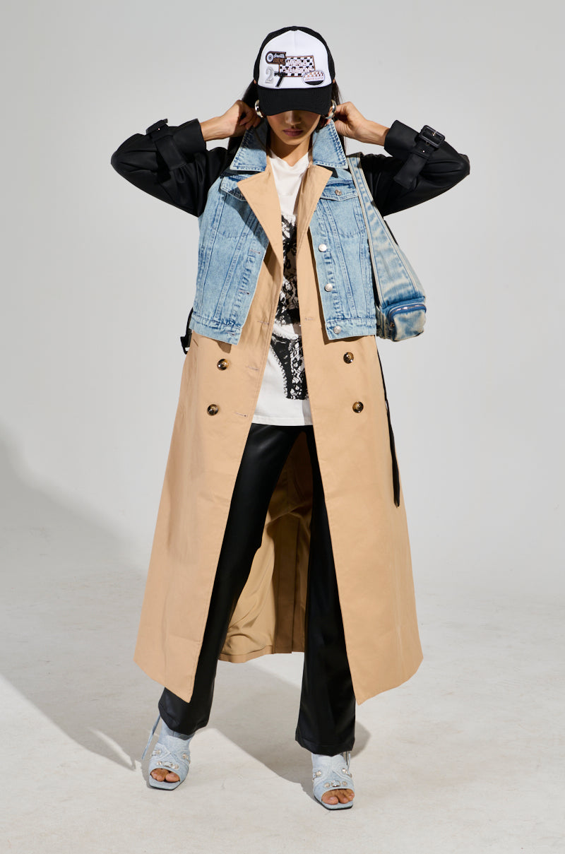 Wearing the CITY SLICKER MIXED MEDIA TRENCH, a person shows off a chic layered look, featuring a denim jacket underneath, paired with a black shirt and graphic tee. They are styled with black pants, blue shoes, and a cap, holding their hands near its brim. The vibrant background enhances the focus on their ensemble.